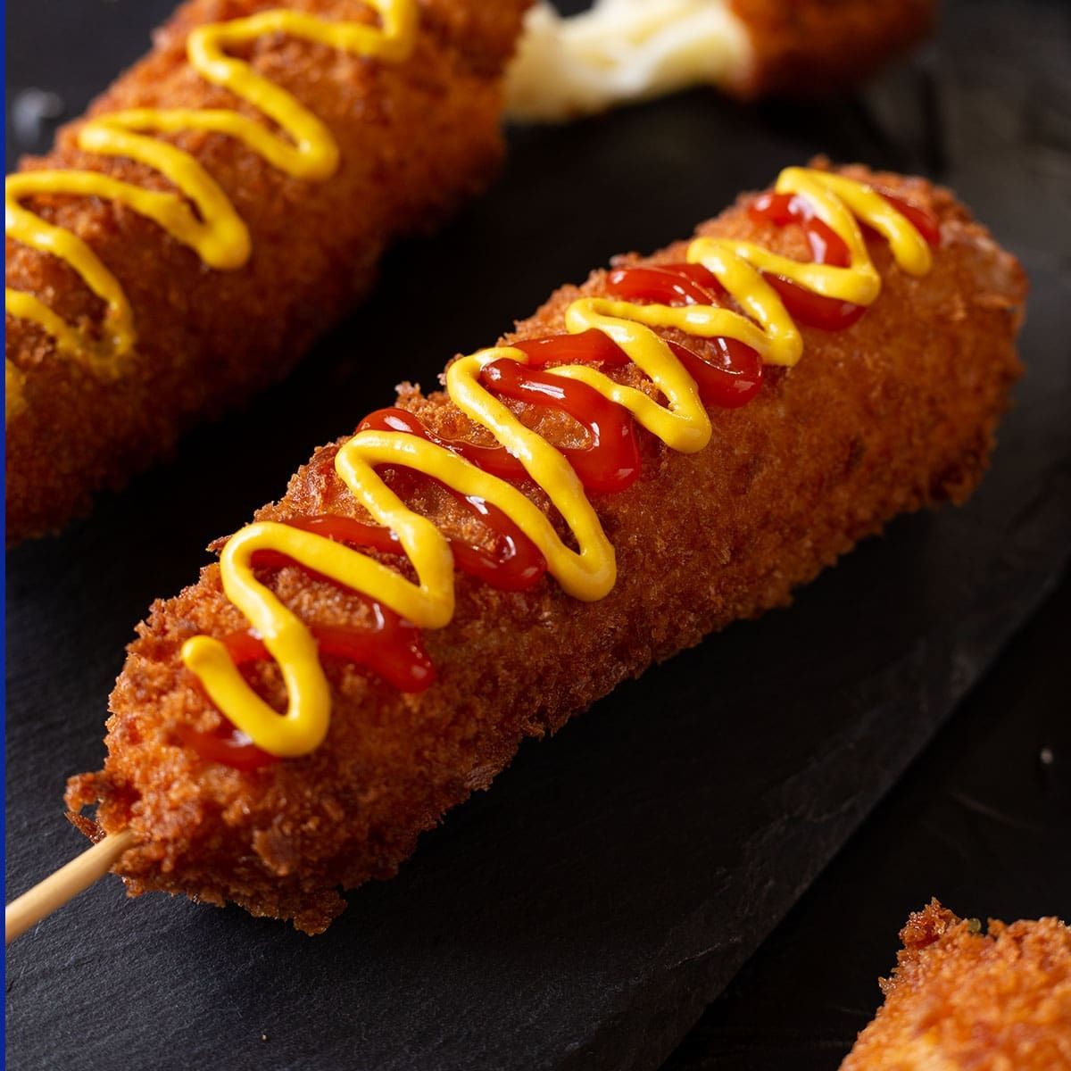 Close-up of a corn dog topped with ketchup and mustard, served on a black slate surface.